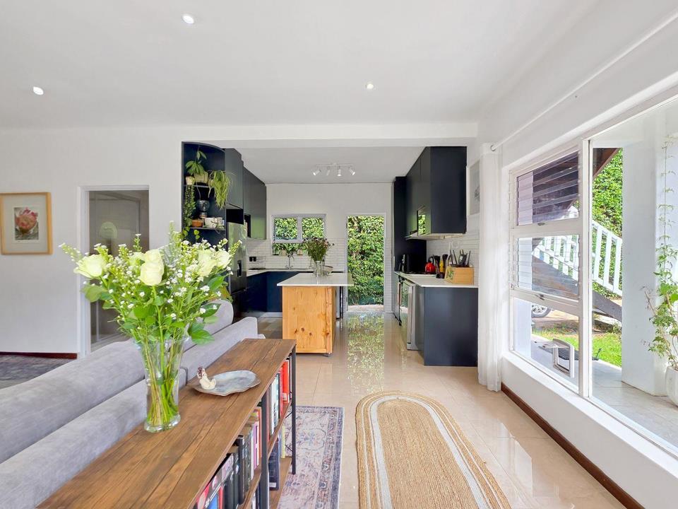 Open-plan lounge viewed from the seating area looking toward the kitchen, with a timber bookshelf coffee table, grey sofa, jute rug, large picture window with garden views, and the navy kitchen cabinetry visible in the b
