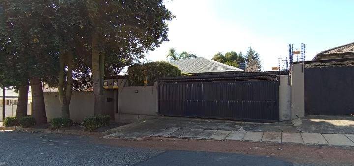 Street-facing exterior of a single-storey Kensington house showing a plastered boundary wall, mature trees, and a wide dark-metal double automated gate with a glimpse of the corrugated-iron pitched roof beyond.