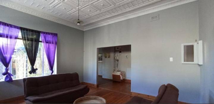 Formal lounge with an ornate white pressed-tin ceiling, dark hardwood timber floors, purple sheer curtains on a large window, two brown leather sofas, and an open archway leading to an adjacent room.