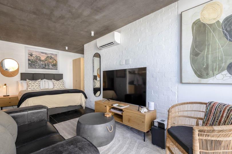 Studio interior showing the sleeping zone with a king-size bed and upholstered grey headboard in the background, the TV and media unit in the centre, and the lounge seating area in the foreground, all beneath an exposed 