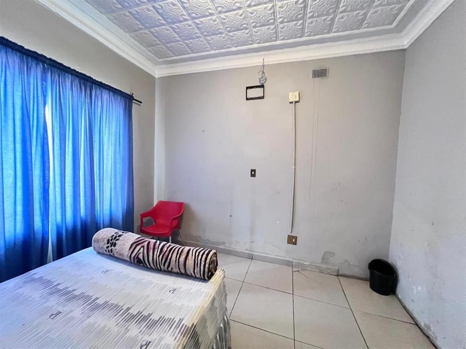 Guest bedroom with white tiled floor, decorative pressed ceiling, blue curtains, a double bed with zebra-print bolster, a red plastic chair, and a wall-mounted TV bracket on grey-painted walls.
