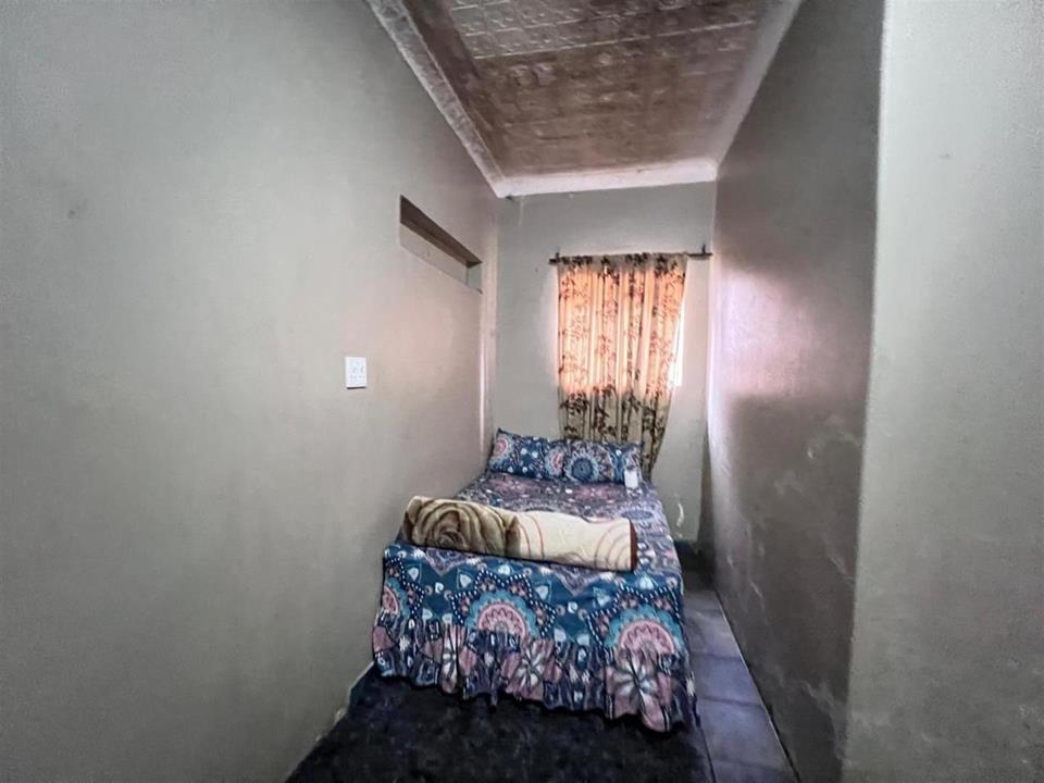 Narrow single guest bedroom with dark carpet, grey plastered walls, decorative pressed ceiling, a single bed with floral bedding, and a small curtained window at the far end.