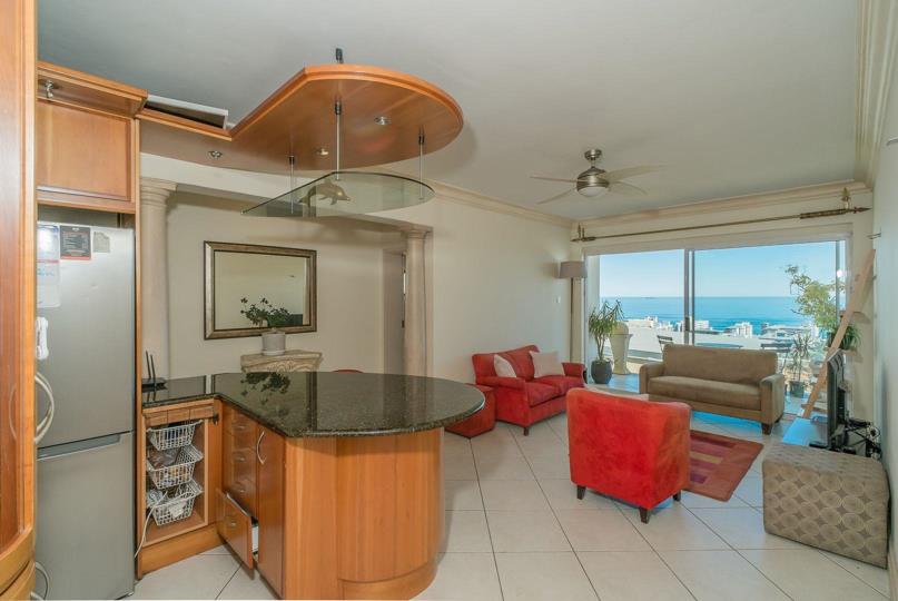 Open-plan kitchen and lounge showing curved timber cabinetry, a dark granite breakfast counter with pendant lighting, stainless-steel fridge, and beyond it a furnished living area with sliding doors opening to an ocean-v