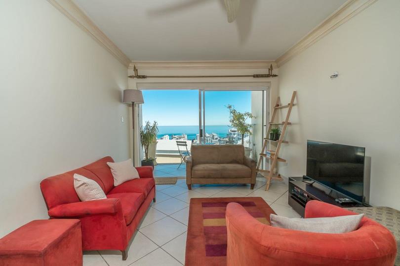 Bright lounge with red sofas, tiled floor, ceiling fan, and large sliding glass doors fully open to a balcony with an unobstructed view of the Atlantic Ocean and Sea Point skyline.
