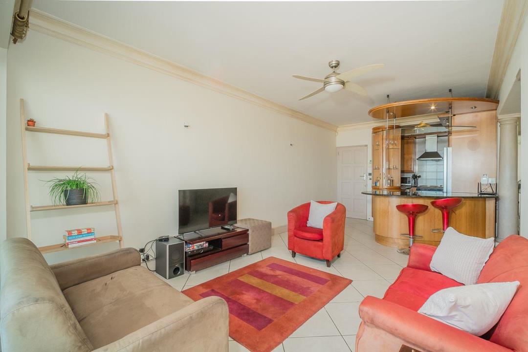 Living room with red and beige sofas, a flat-screen TV on a media unit, ceiling fan, decorative ladder shelf, and a curved timber kitchen bar with red bar stools visible in the background.