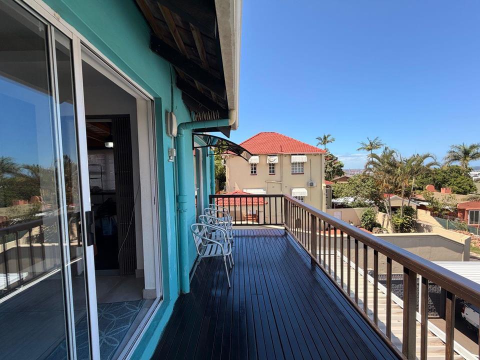 Upper-level balcony of a teal-painted house with dark composite decking, metal railing, two white metal chairs, and a sliding glass door opening to the interior lounge; neighbouring red-tiled rooftops and palm trees visi