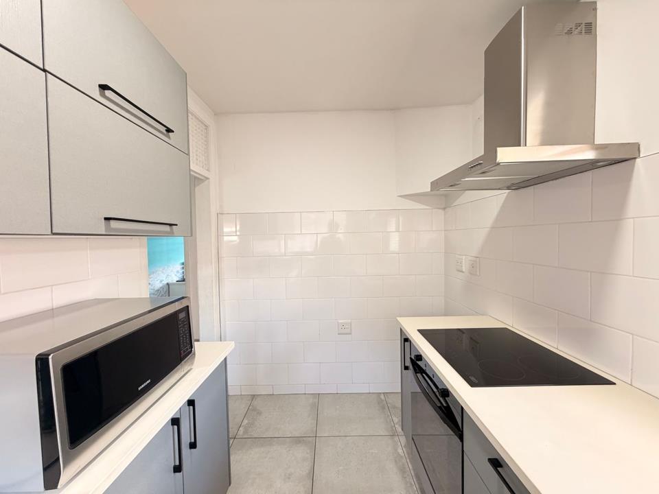 Second angle of the renovated kitchen showing grey flat-panel upper and lower cabinets with black handles, white subway-tile splashback, black ceramic hob with stainless-steel extractor hood, Samsung microwave, and light