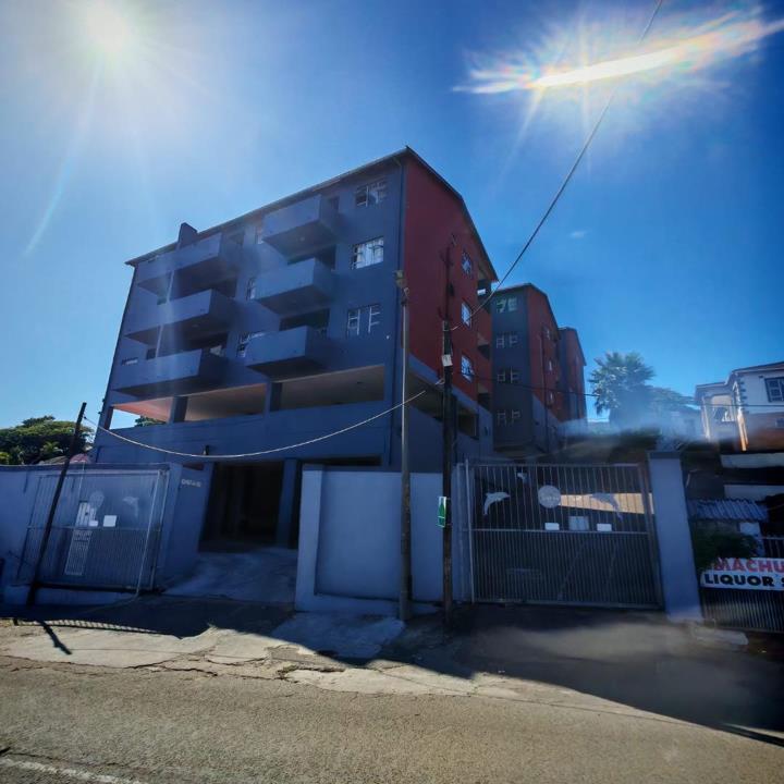 Street-level view of a four-storey apartment block with a blue and red painted exterior, gated vehicle entrance on the left, and a liquor store visible on the adjacent property to the right, photographed under bright mid