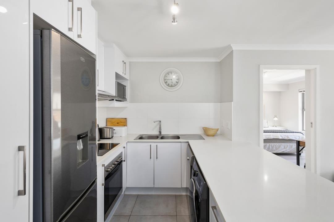 Close-up view of the white open-plan kitchen showing a large quartz island counter in the foreground, stainless-steel double-door fridge-freezer with water dispenser on the left, built-in oven and hob, microwave on an up
