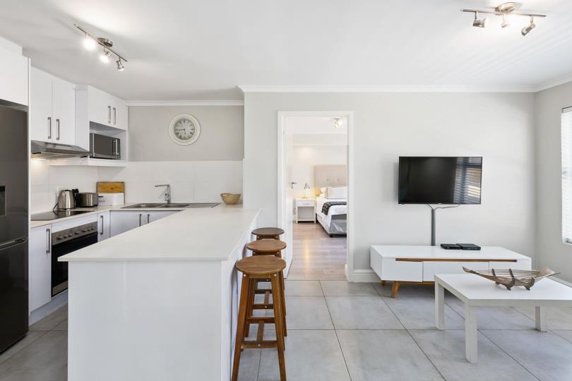 Alternative angle of the open-plan kitchen and living room, highlighting the white quartz breakfast bar with three timber stools, kitchen appliances, flat-screen TV on white TV stand, white coffee table, and blue accent 