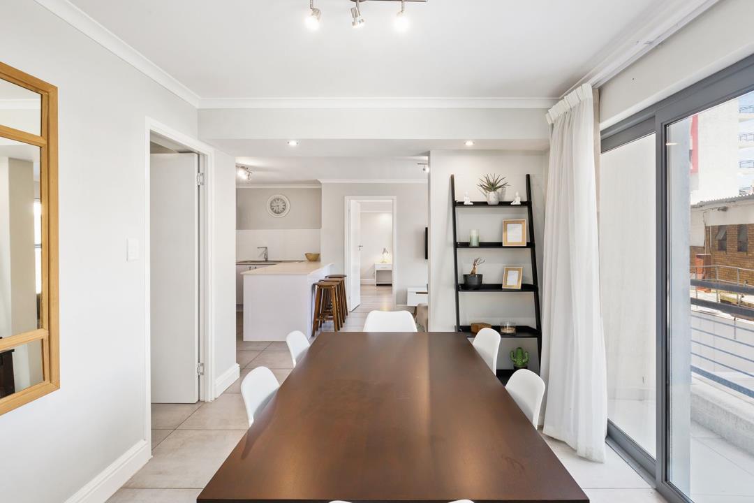 Dining room with a large dark-timber six-seater dining table and white chairs, a lean-to display shelf with décor, sliding glass door with white curtains opening to a balcony, and an open view through to the kitchen and 
