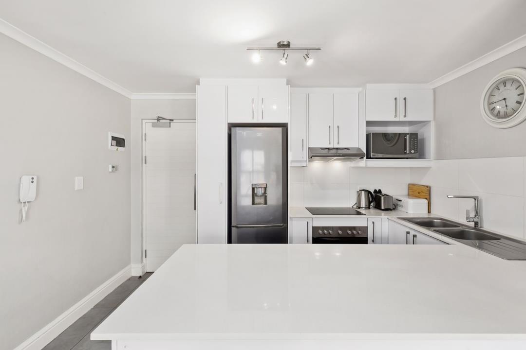 Reverse-angle kitchen shot showing full run of white gloss upper and lower cabinetry, tall fridge-freezer with water dispenser, electric ceramic hob and built-in oven, stainless-steel microwave, kettle and toaster on the