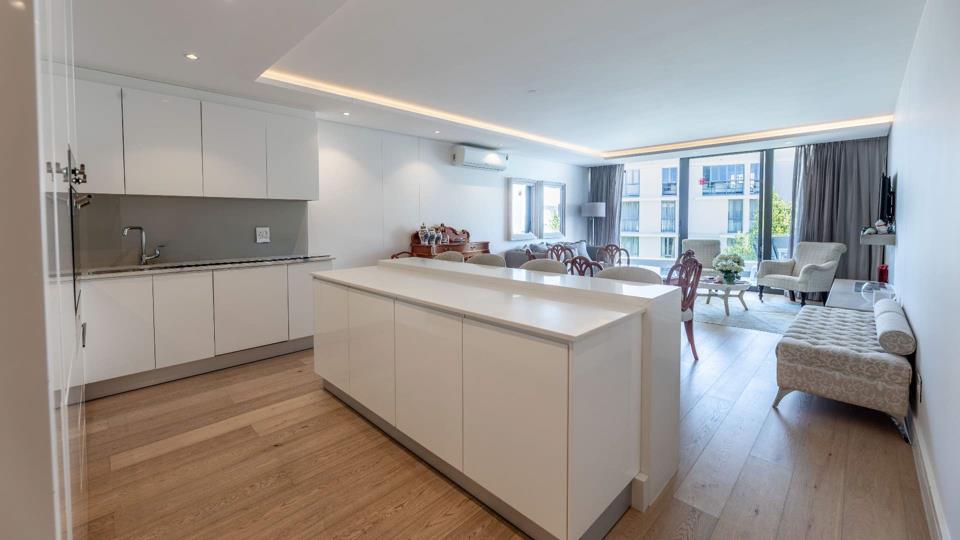 Wide-angle view of the open-plan kitchen and living area, showing the white island bench in the foreground, the full kitchen run along one wall, and the furnished lounge with floor-to-ceiling glazing and a balcony beyond